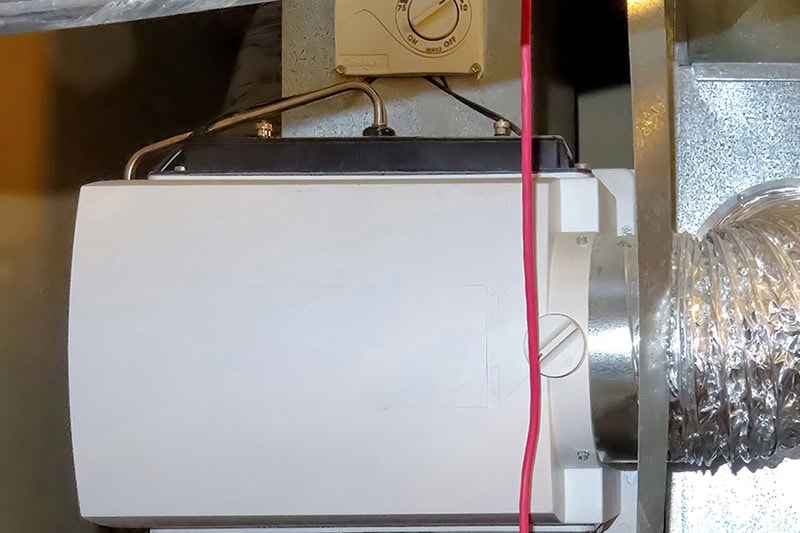 A close-up view of a whole-house humidifier mounted on an HVAC duct, featuring a humidistat control dial and an attached flexible duct hose.